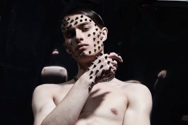 Sanat ve moda. Çıplak gövdeli yakışıklı bir esmer adam yüzünde ve elinde metal çivilerle siyah bir arka planda poz veriyor.. 