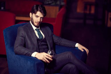 Lüks dairelerde bir bardak şarapla koltukta dinlenen zarif klasik takım elbiseli yakışıklı bir esmer adam portresi. Erkek güzelliği, moda. İş biçimi.