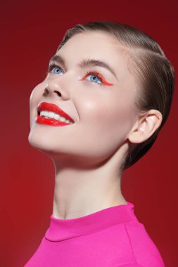 Kozmetik ve makyaj malzemeleri. Parlak kırmızı rujlu ve gözlerinde kırmızı oklar olan sevimli sarışın bir kız büyüleyici bir şekilde gülümsüyor ve kırmızı bir arka plana bakıyor. Güzellik konsepti.