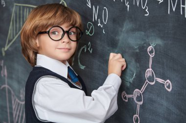 Zeki çocuklar. Eğitim. Düzgün okul üniforması ve gözlükleriyle akıllı, gülümseyen bir çocuk tahtaya oturmuş kimyasal formüller ve şemalar yazıyor..  
