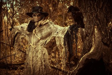 A terrible Scarecrow with a canvas sack on his head and in a burlap cloth stands in the dark thicket of the forest with a black raven on his hand. Vintage style. Halloween Tales. Horror, thriller. clipart