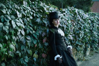 A beautiful feminine young lady in an elegant black suit of the 19th century standing against the backdrop of green foliage.  clipart