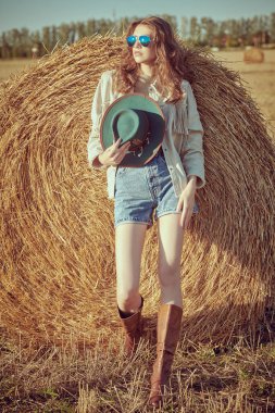 Modern hippi ve vahşi batı tarzı. Güzel manken kız, batan güneşin altında samanlıkta samanlığın yanında kot elbise, şapka ve deri botlarla poz veriyor. Yaz ve sonbahar modası.