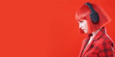 Güzellik, moda. Kırmızı dudaklı, kızıl saçlı, kırmızı kareli ceket ve kulaklık takan şık bir kızın yan görüntüsü. Fotokopi alanı olan kırmızı stüdyo. DJ, parti kızı.