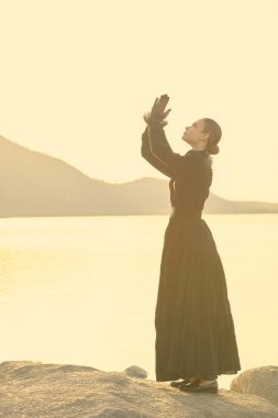 Zarif, siyah, 19. yüzyıl elbisesi içinde genç bir kadın gün batımında denizin kenarında duruyor. Dua etmek için ellerini birleştiriyor ve umutla bakıyor. 19. yüzyıl romanı.