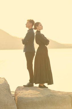 Güzel bir kadın ve deniz kenarında kollarını kavuşturmuş birbirlerine sırt çeviren yakışıklı bir genç adamın yan görüntüsü. Sevgi dolu bir ilişki. Drama, aşk romanı. 