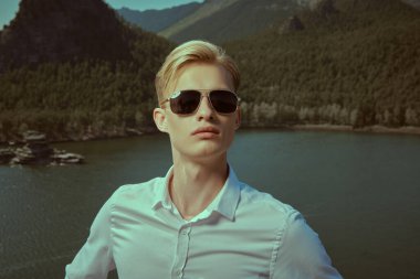 Beyaz gömlekli ve siyah güneş gözlüklü yakışıklı ve kasıtlı bir adam bir dağ manzarasına karşı duruyor ve kameraya bakıyor. İş konsepti, hedef belirleme ve başarı. Tatil ve seyahat.