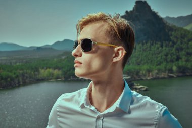 Beyaz gömlekli, kasıtlı ve siyah güneş gözlüklü yakışıklı bir adam bir dağ manzarasına karşı duruyor ve uzaklığa bakıyor. İş konsepti, hedef belirleme ve başarı. Tatil ve seyahat.