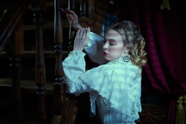 Beyaz dantelli elbiseli zarif, güzel bir aristokrat kadın geceleri merdivenin yanındaki klasik bir dairede duruyor. Viktorya tarzı. Tarihi roman..