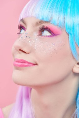 Anime makyajı. Parlak makyajlı, parlak çilli ve pembe arka planda kurnazca gülümseyen menekşe mavisi peruklu güzel kız. Saç stili, saç boyası, makyaj. Japon animasyon tarzı. 