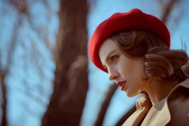 Yağmurluk giymiş, kırmızı bereli güzel bir kızın parkta dikilip düşünceli bakışlarının yan görüntüsü. Bahar ve sonbahar modası.