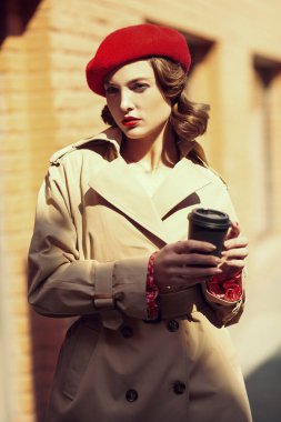 Trençkotlu ve kırmızı bereli güzel, zarif bir kadının portresi elinde bir fincan kahveyle bir şehir caddesinde yürüyor. Güzellik, moda. Zarif retro tarzı.