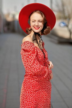 Açık kırmızı elbiseli, kırmızı şapkalı, zarif güneş gözlüklü, güzel güneşli bir günde şehrin caddesinde neşeli bir şekilde gülümseyen çekici bir kız. Kadınlık ve eski tarz.