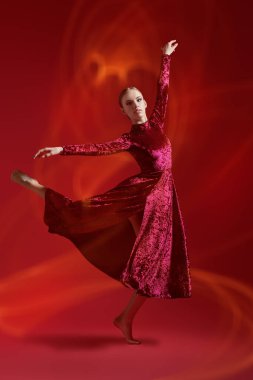 Kırmızı kadife elbiseli zarif bir dansçı kız stüdyoda kırmızı arka planda dans ediyor. Tam boy portre.
