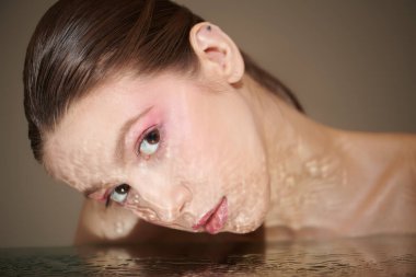 Makyajlı güzel bir kız, kameraya sakince bakar ve yüzünü temiz suyun üzerine eğer. Stüdyo çekimi. Kadın güzelliği ve sağlığı. Kozmetik ve cilt bakımı. 