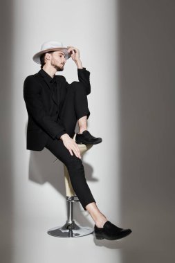 Zarif bir esmer adam portresi şık siyah bir takım elbise ve beyaz bir şapka içinde beyaz bir duvarın kenarında ışık huzmesi ile aydınlatılmış bir taburede oturuyor. Moda fotoğrafı. Işık ve gölge.
