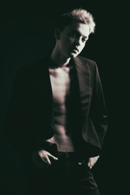 Siyah smokinli yakışıklı bir genç adam portresi çıplak bir bedenin üzerinde, gölgede karanlık bir arka planda poz veriyor. Erkek modası ve güzellik. 