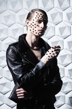 Agresif bir moda. Siyah deri ceketli, yüzünde ve elinde metal sivri uçlu beyaz bir arka planda poz veren vahşi bir gencin portresi..