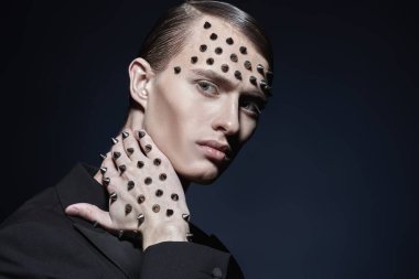 Moda fotoğrafı. Siyah kuyruklu yakışıklı, vahşi bir adamın portresi. Yüzünde ve elinde siyah bir arka planda metal çiviler var.. 