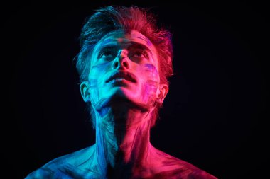 Renkli boyalı yüzü ve vücudu olan sakin, yakışıklı bir adam neon mavisi ve pembe bir ışıkla siyah bir arka planda dikiliyor. Sanat ve moda. Vücut boyası.