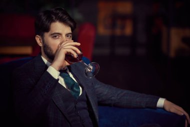 Şık, klasik takım elbiseli yakışıklı bir adamın portresi lüks dairelerde bir bardak şarapla kanepede dinleniyor. Erkek güzelliği, moda. İş biçimi.
