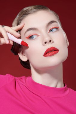 Makyajda kırmızı renk. Parlak makyajlı güzel bir kız modelin portresi. Elinde muhteşem kırmızı bir ruj tüpü var. Burgonya arkaplanı. Güzel kız.. 
