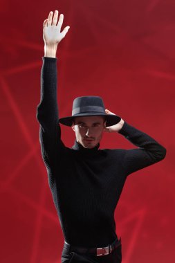 Yakışıklı, şık, siyah günlük giysiler içinde zarif bir genç adam ve koyu kırmızı bir arka planda koluyla şapka pozu veriyor. Erkek modası ve güzelliği. 