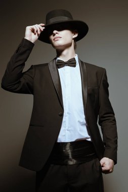Siyah smokinli yakışıklı bir genç. Papyonlu ve şapkalı. Gri stüdyo arka planında gölgesi var. Erkek modası ve güzelliği. 