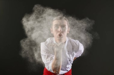 Profesyonel erkek pasta şefi nefis bir yemek hazırlar ve avucundan un üfler. Un, karanlık bir stüdyo arka planında havada uçuşuyor. Yemek ve yemek..