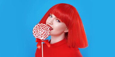 Parlak kırmızı dudakları ve kızıl omuz hizasında saçları olan güzel, zarif bir kız lolipopu neşeyle yalıyor. Mavi stüdyo geçmişi. Güzellik, kozmetik, makyaj. Saç stili, saç boyası.