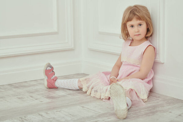 Красивая 4-летняя девочка с светлыми волосами, одетая в розовое летнее платье сидит спокойно прислонившись к стене в белой классической квартире. Дети, детство и мода.