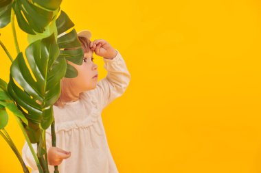 Etraftaki dünyayı keşfediyorum. Palmiye yapraklarının yanında duran komik küçük bir kız ilgiyle ona bakıyor. Turizm, yaz tatili. Sarı arka plan. Boşluğu kopyala.