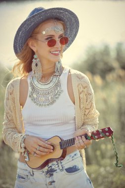 Güneşli bir yaz gününde Boho tarzı kıyafetler ve takılar içinde neşeli bir kız açık havada ukulele çalar. Yaz bohem ve modern hippi tarzı. Yaz tatili.