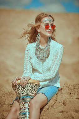Boho elbiseli ve mücevherli mutlu sarışın kız sahilde oturur ve tam-tam davul çalar. Hippi ve boho stili. Yaz tatili. Serbest yaşam biçimi.      