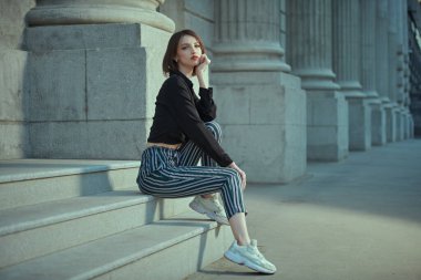 Siyah bluzlu ve çizgili pantolonlu, kısa saçlı güzel esmer bir kızın şehir caddesindeki bir binanın merdivenlerinde oturduğu uzun bir kare. Moda ve tarz. Kaygısız yaz günü.