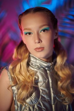 Dalgalı sarı saçları ve turkuaz göz makyajıyla tropik bitki arka planında gümüş desenli ceketiyle poz veren güzel bir kızın portresi. Güzellik, makyaj ve saç stili. Yüksek moda.