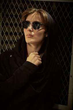 Siyah kapüşonlu hırka ve siyah güneş gözlüğü takan genç bir çocuk metal bir ağ çitinin yanındaki arka sokakta duruyor. Ergenlik, modern genç nesil ve yaşam tarzı.