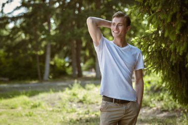 Mutlu bir genç adam yeşil bir yaz parkında duruyor ve gülümsüyor ve neşeli bir şekilde uzaklaşıyor. Mutlu insanlar, olumlu duygular, yaşam tarzı. 