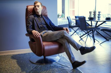 Yakışıklı genç işadamı modern bir ofiste deri koltukta oturup düşünceli bir şekilde uzaklara bakar. Saygıdeğer bir ofis ve iç mekan. İş konsepti, başarı.