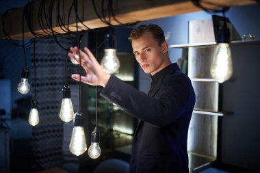 Zeki, bakımlı genç bir adam modern dairesinde şık lambaların yanında duruyor. Ev ve ofis için ev ve mobilyalar.