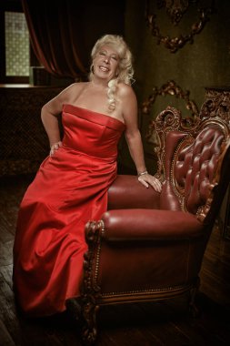 Retro tarzı. Zarif, kırmızı bir gece elbisesi içinde yaşlı ve güzel bir bayan klasik iç mimarisi olan bir odada eski bir koltuğa yaslanıyor. Lüks. Yaşlı Kontes..