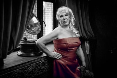 Kırmızı bir gece elbisesi içinde zarif yaşlı bir kadın bir patifonun yanında duruyor ve klasik bir iç mimariye sahip bir odada gülümsüyor. Retro tarzı. Güzellik ve ihtiyarlık.