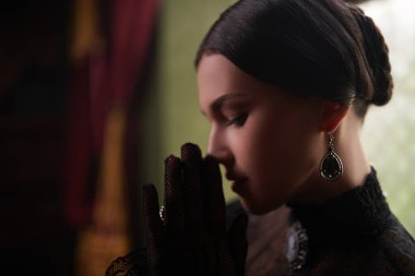 Asilzade kökenli sofistike bir esmer kız, elleri duada katlanmış, koyu siyah bir elbise içinde duruyor. Profilde bir portre. Romandaki kadın kahraman, dramatik hikaye..