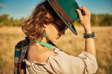 Boho tarzı kıyafetleri ve aksesuarları olan muhteşem bir kız batan güneşin parlak ışıklarıyla bir tarlanın ve mavi gökyüzünün arka planında duruyor. Yaz tatili, seyahat. Bohem, modern hippi tarzı.. 