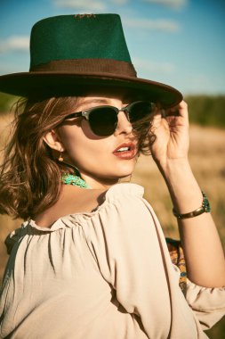 Tarlada ve mavi gökyüzünde kadınsı bir bluz, şapka ve güneş gözlüğü takan çekici bir kız. Yaz tatili, seyahat. Bohem, modern hippi tarzı.. 