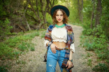 Modern hippi tarzı. Yeşil bir yaz parkında şık giyinmiş çekici bir boho kızı poz veriyor. Yaz tatili, seyahat. Denim modası.