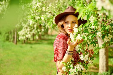 Bahar sezonu. Hippi tarzı kıyafetleri ve aksesuarları olan güzel bir kız parkta çiçek açan ağaçlardan hoşlanır. Boho ve hippi tarzı. Güzellik ve moda. Boşluğu kopyala.