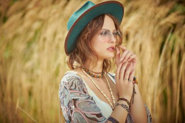 Boho tarzı kıyafetler ve aksesuarlarla güzel romantik bir kız sazlıkların yanındaki bir tarlada duruyor ve uzaklığa bakıyor. Boho ve hippi tarzı. Yaz, tatil.