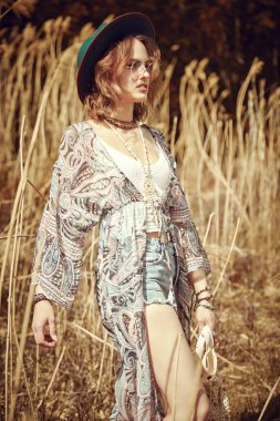 Boho tarzı kıyafetleri ve aksesuarları olan şık, güzel bir kız sazlıkların yakınındaki bir tarlada dikilip mesafeye bakıyor. Boho ve hippi tarzı. Tatil, seyahat. 