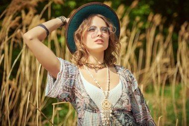 Boho tarzı kıyafetler ve aksesuarlarla güzel romantik bir kız sazlıkların yanındaki bir tarlada duruyor ve uzaklığa bakıyor. Boho ve hippi tarzı. Yaz, tatil.